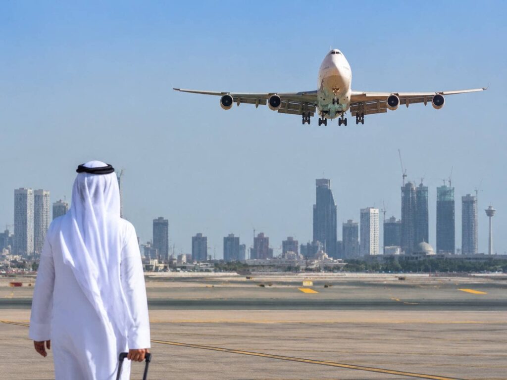 Man in a white kandura standing on an airport tarmac with a suitcase, watching a large airplane approach for landing against a city skyline.