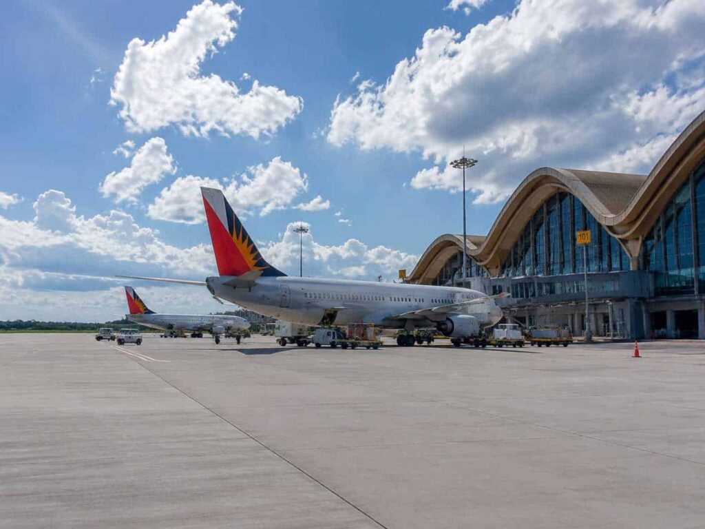 Airplanes parked at a sunny airport tarmac with a curved-roof terminal in the background