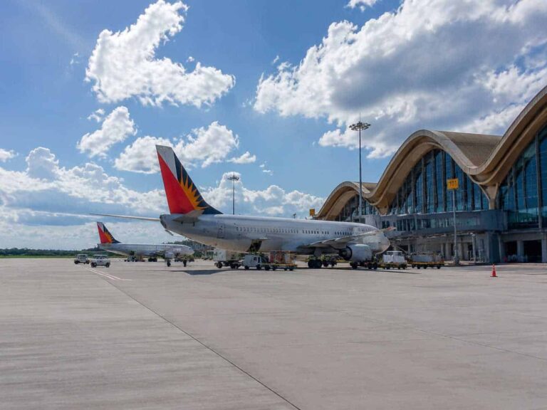 Airplanes parked at a sunny airport tarmac with a curved-roof terminal in the background