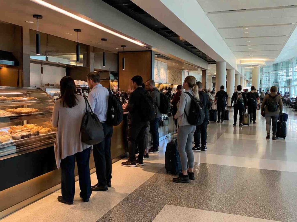 Travelers line up at a pastry counter in a busy airport terminal, with display cases of baked goods on the left and passengers with luggage waiting in line.",