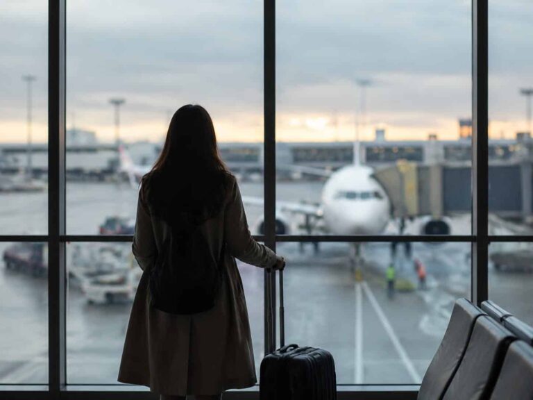 Woman with a rolling suitcase stands at an airport gate, gazing at a parked airplane through large windows at dusk.