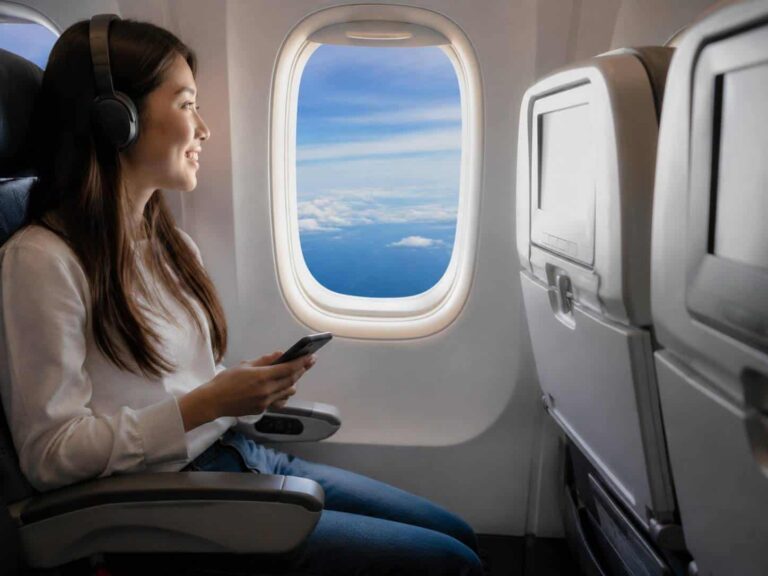 Woman wearing over-ear headphones seated on an airplane, using a smartphone by the window.