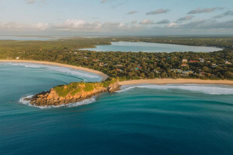Aerial view of a rocky headland extending into turquoise water, with a sandy beach and a tree-lined residential area along the shore.