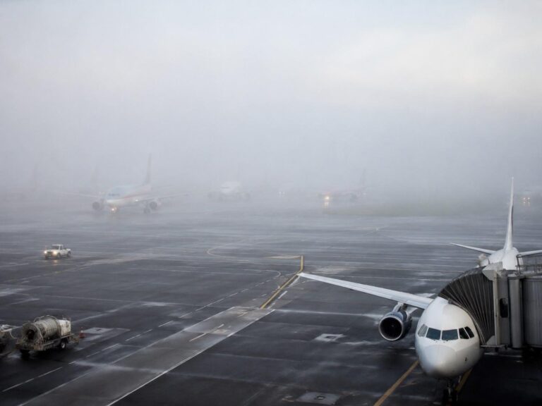 Airport gate with a jet bridge attached to a white airplane on the right; fog obscures distant aircraft and tarmac is wet from rain/fog.