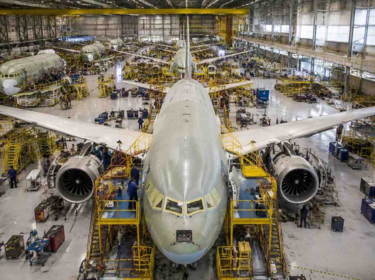 Wide view of a large airliner in a maintenance hangar, surrounded by yellow scaffolding and technicians around the engines.