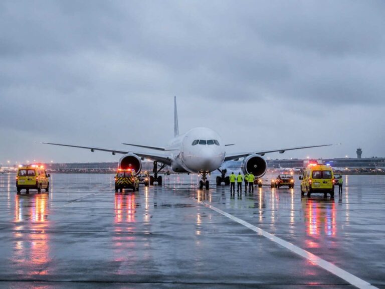 Front view of a large white airliner on a wet runway, with emergency vehicles and ground crew in high-visibility jackets.