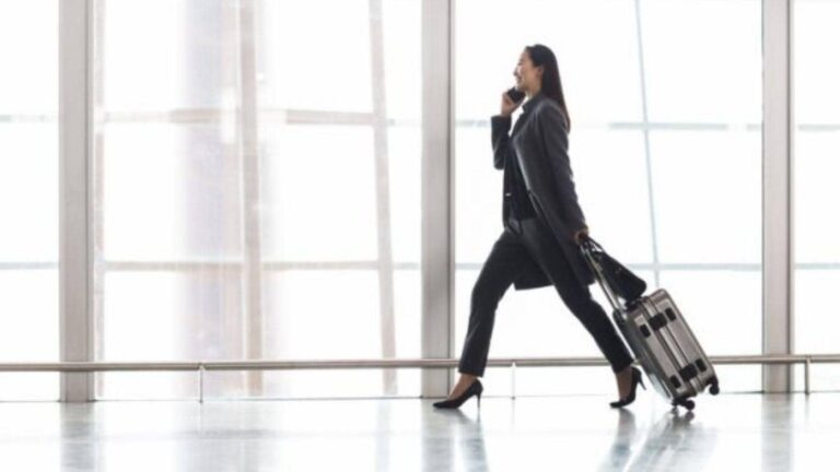 Professional woman in a blazer walks through an airport with a rolling suitcase and high heels, talking on her phone.