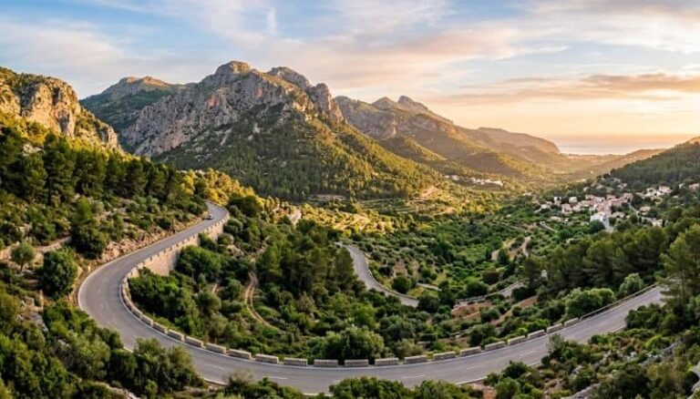 Winding mountain road curves through green hills at sunset, with rocky peaks and a distant valley village.