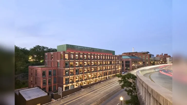 Brick building with many windows beside an elevated highway at dusk, urban street scene with light trails from cars.