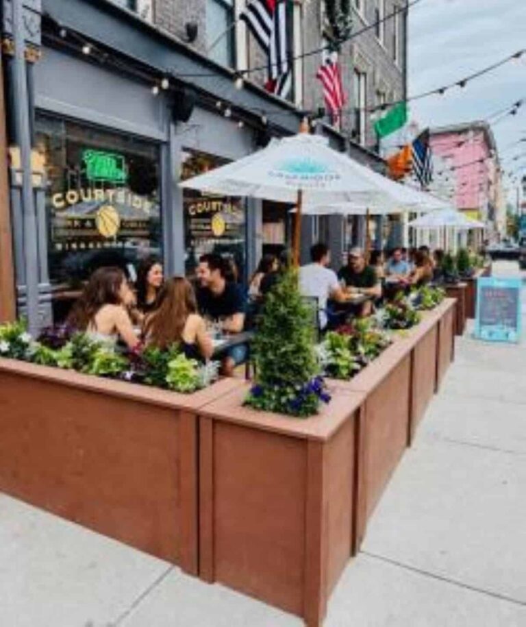 Outdoor sidewalk cafe with groups of people dining under white umbrellas, bordered by wooden planters with green plants.