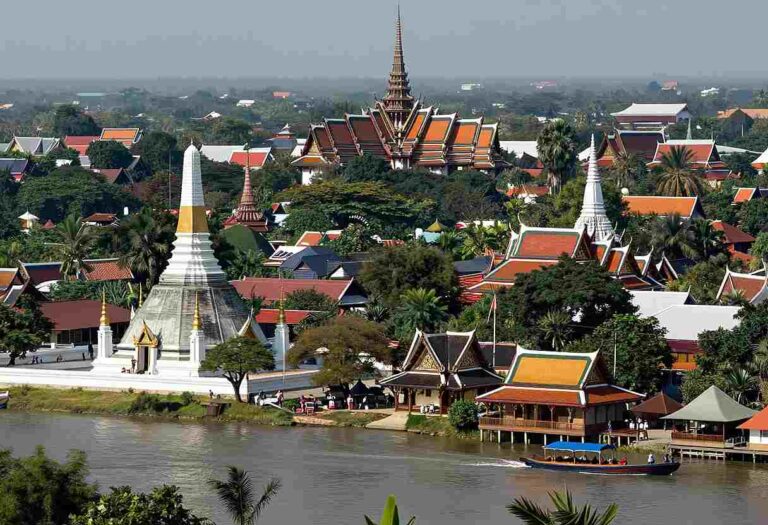 Aerial view of a Thai temple complex by a river, with a white stupa, colorful tiered roofs, and lush trees nearby.