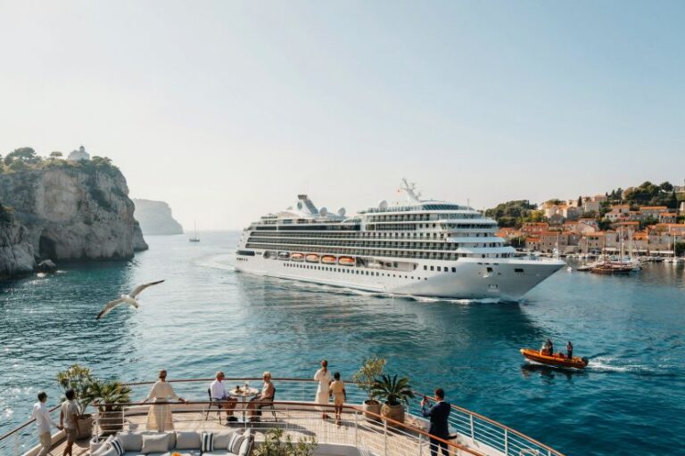 A large cruise ship sails into a calm harbor near a coastal town, with people on a yacht deck watching the scene.