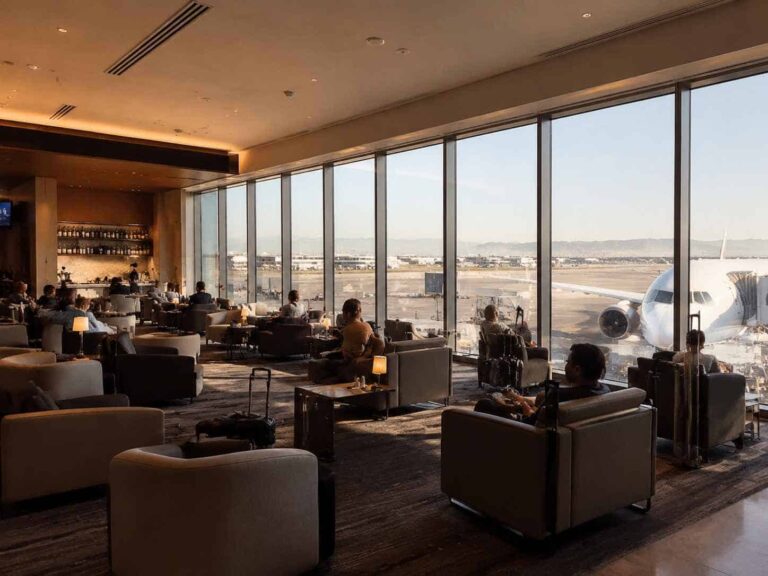 Passengers lounge in a spacious airport lounge with floor-to-ceiling windows overlooking the tarmac and a parked aircraft outside.