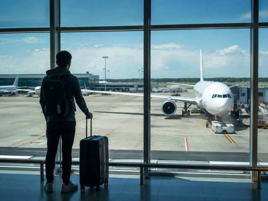 Traveler with a backpack and rolling suitcase at the airport, looking at a plane at the gate outside the window.”](sic)