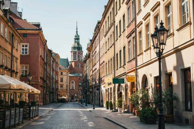 Sunlit cobblestone street in a historic European town, with colorful facades and a distant church tower.