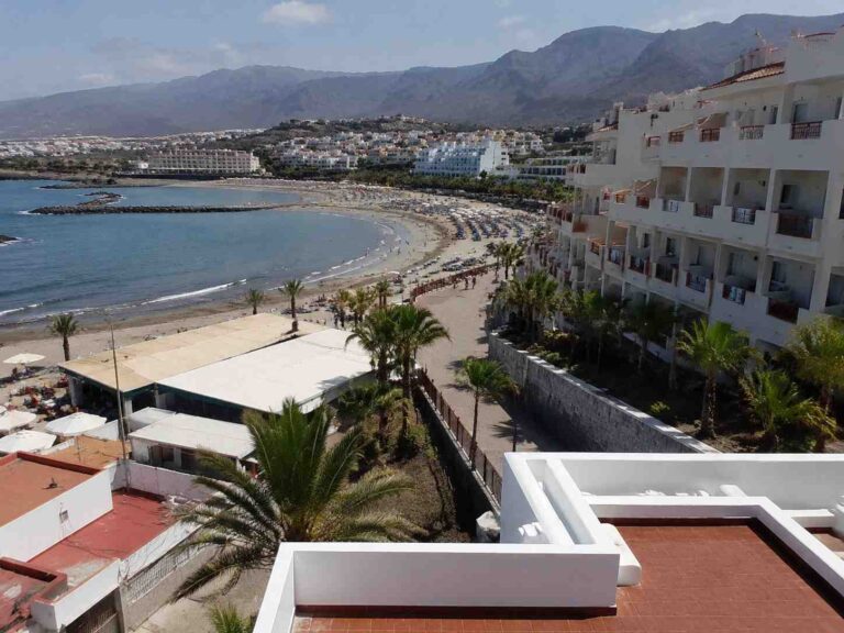 Coastal resort with a sandy beach, palm trees, and white apartment buildings along the shore, mountains in the distance