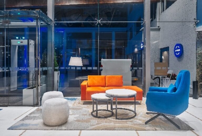 Modern hotel lobby lounge with bright orange sofa, blue accent chair, white round tables, and a light gray pouf on a patterned rug.