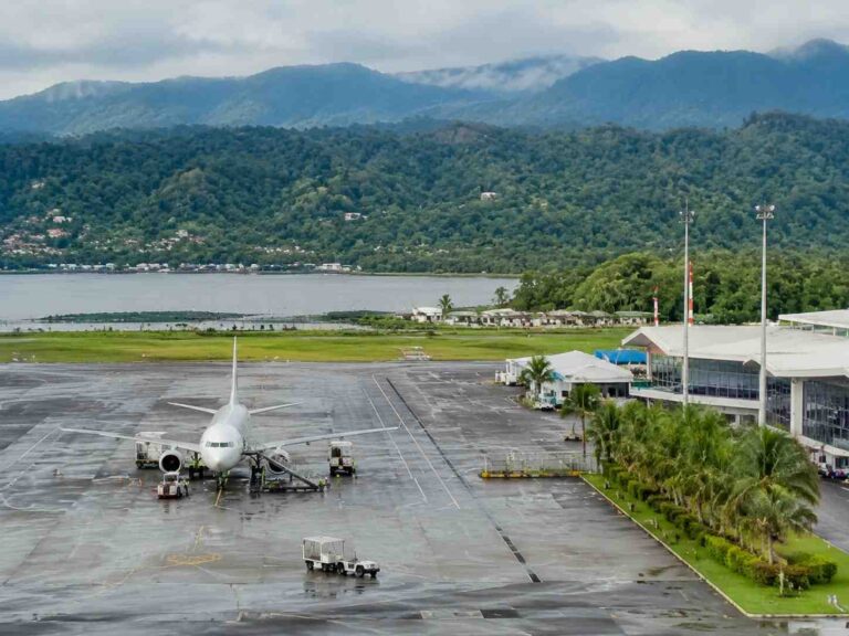 Airplane parked at a gate on a wet tarmac with a terminal building and green hills in the distance