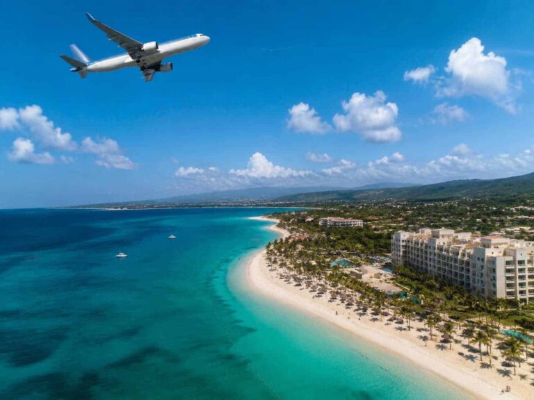 White commercial airplane flying over a tropical beach resort with turquoise water, white sand, and palm-lined shoreline.