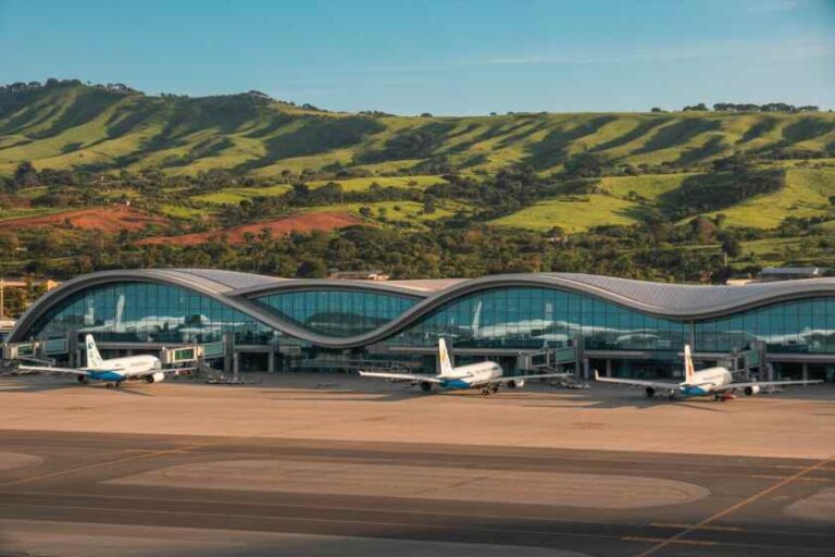 Airplanes parked at gates in front of a glass-fronted, wavy-roof airport terminal with green hills in the background.