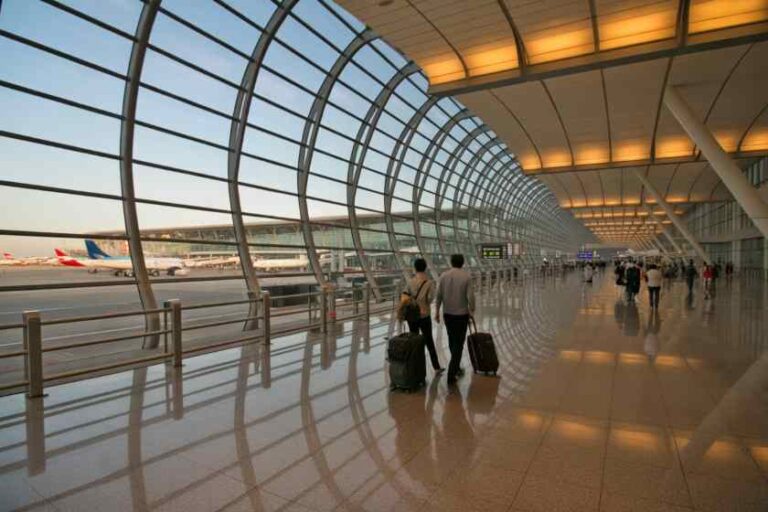 Interior of a modern airport terminal with curved glass walls and travelers pulling suitcases along a polished floor outside planes are visible at gate side.