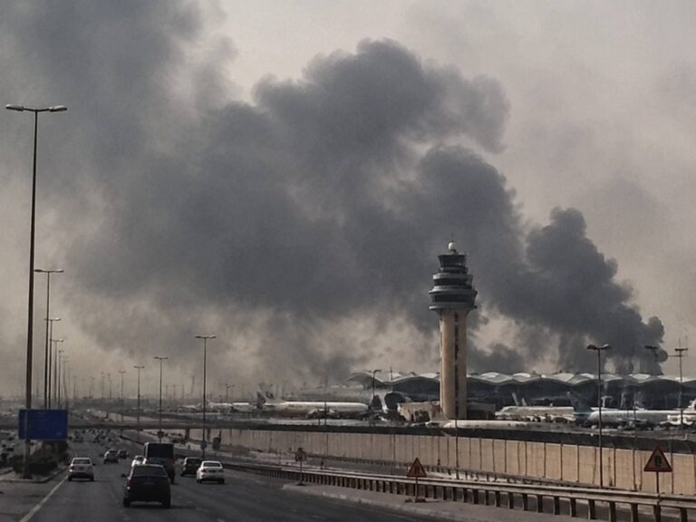 Airport view with control tower beside a runway and a large dark smoke plume rising behind the terminal area, suggesting a fire nearby.