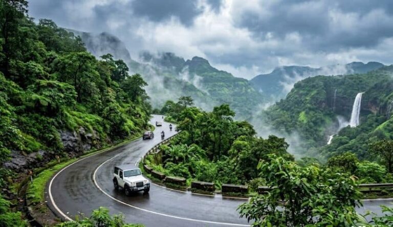 Winding mountain road through lush green forest with mist and a distant waterfall in the background, several cars and motorcycles on the curve in wet conditions from rain.