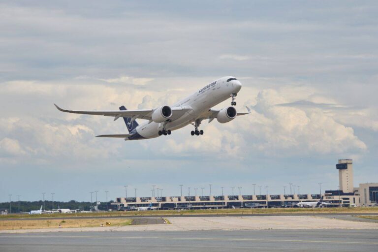 White commercial jet taking off from a runway, gear still down, with airport terminals in the background under a cloudy sky.