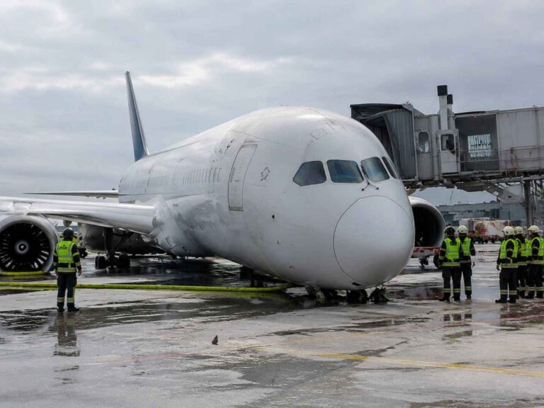 Wide-body airliner at a jet bridge with ground crew in high-visibility vests on a wet tarmac.