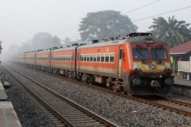 Orange passenger train at a rural station on double tracks with palm trees and a platform in the background.