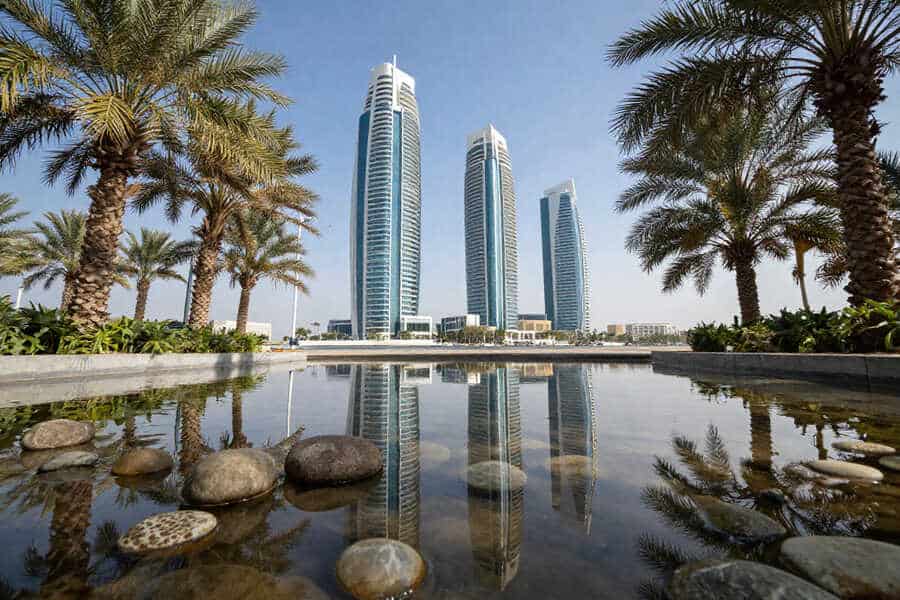 Modern city skyline with three tall glass towers, palm trees, and a reflective pool in the foreground.