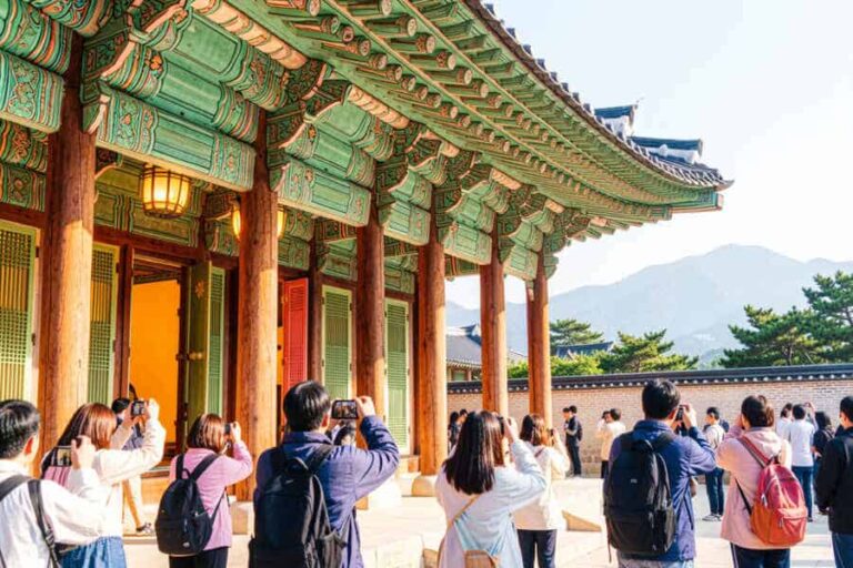 Tourists photograph a traditional green-eaved temple with wooden columns and a mountain backdrop.