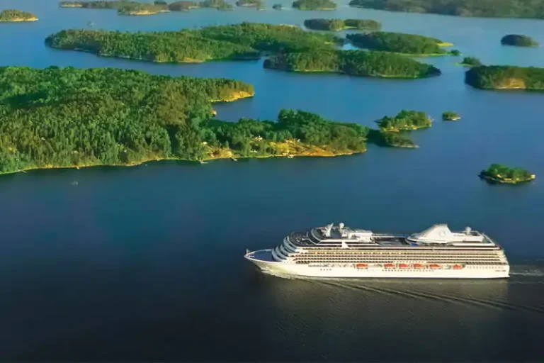 Aerial view of a large white cruise ship sailing through blue water among green forested islands.