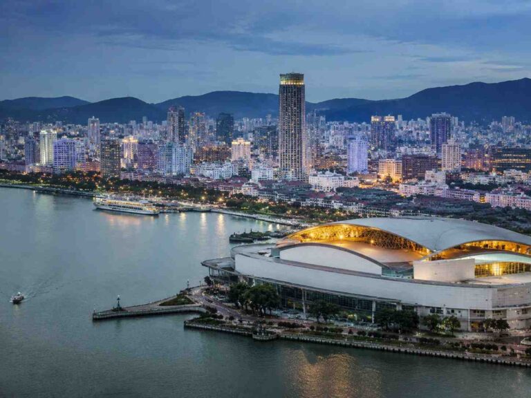 Aerial view of a coastal city at dusk with a curved white-roofed venue in the foreground and a lit skyline beyond.