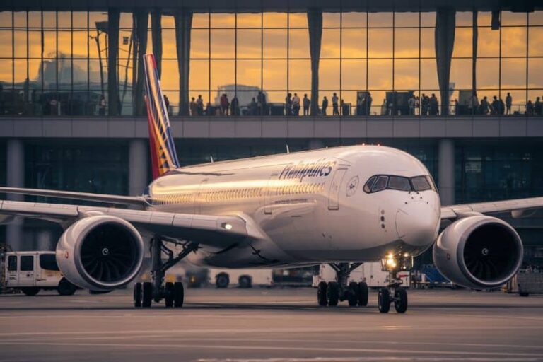 Front view of a large white passenger jet on the runway with a glass-walled terminal in the background.