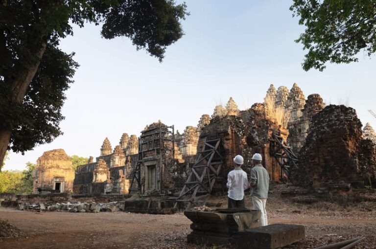 Ancient temple ruins under restoration with wooden scaffolding; two people wearing hard hats stand nearby.