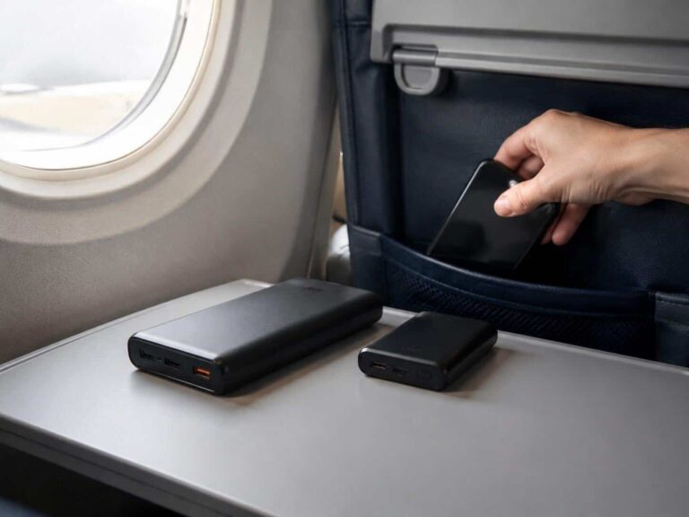Two portable power banks sit on an airplane tray table as a hand holds a smartphone nearby, ready to charge.