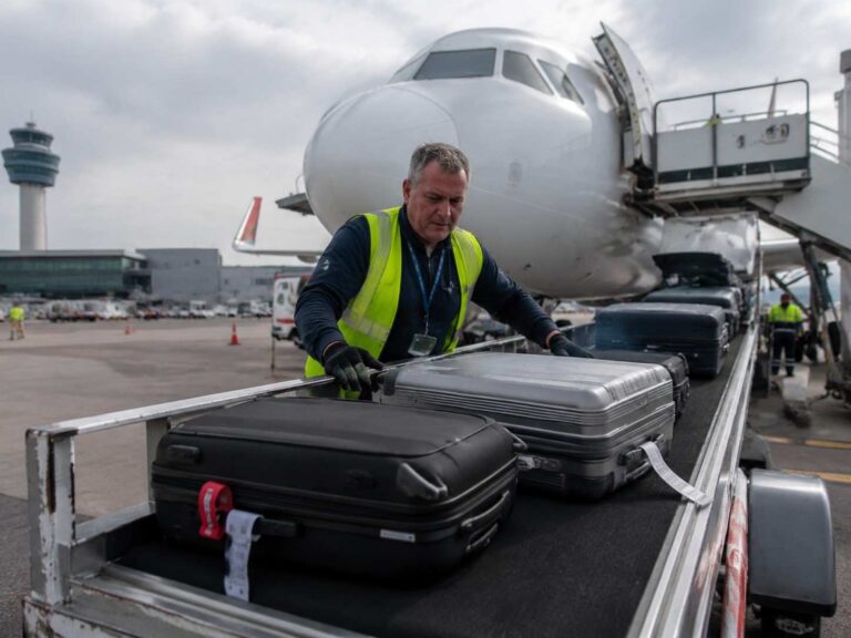 Ground crew member in a hi-vis vest loads black and silver suitcases onto a baggage conveyor beside a parked airplane at the terminal pool of tarmac.
