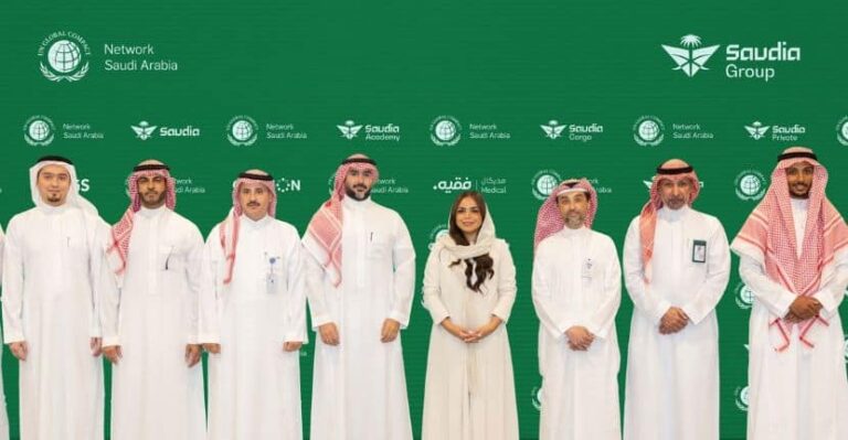 Group of eight people in traditional Saudi attire posing in front of a green Saudia backdrop with logos and branding.