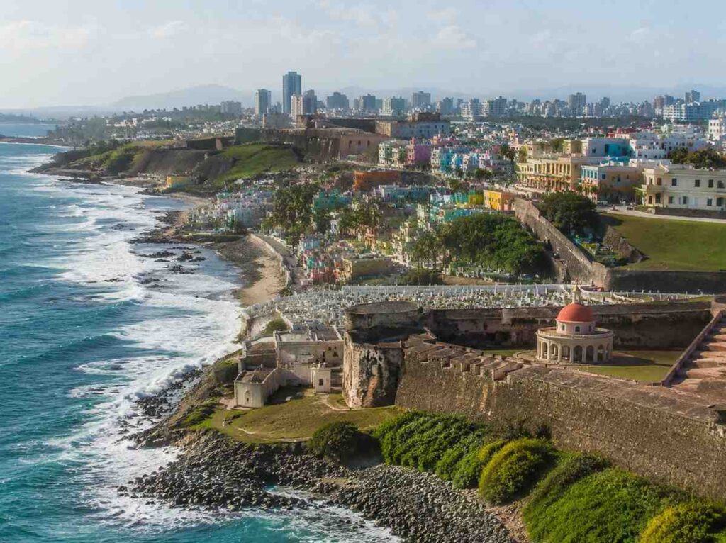 Aerial view of a historic fortress along a rocky coastline, with a colorful city stretching behind and the ocean on the left.