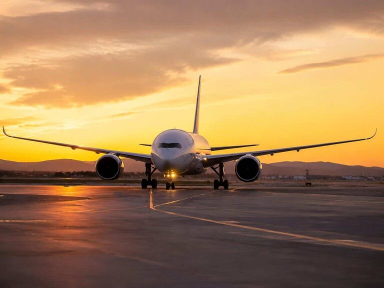 Front view of a large commercial airplane taxiing on a sunlit runway at sunset with an orange sky and distant hills in the background