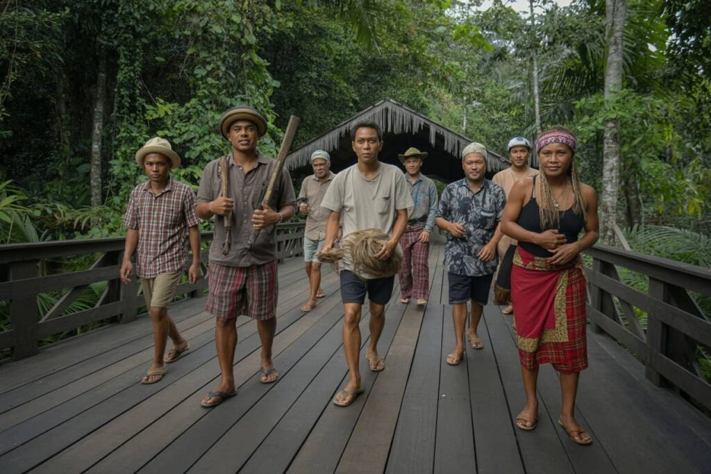 Group of people walking along a wooden boardwalk through a dense tropical jungle, some carrying tools and a woven bundle.