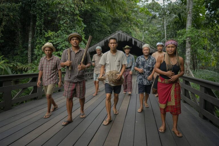 Group of people walking along a wooden boardwalk through a dense tropical jungle, some carrying tools and a woven bundle.