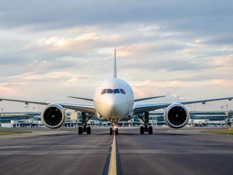 Front view of a large white commercial airliner on a runway, engines visible on both wings, airport terminal in the background under a cloudy sky.