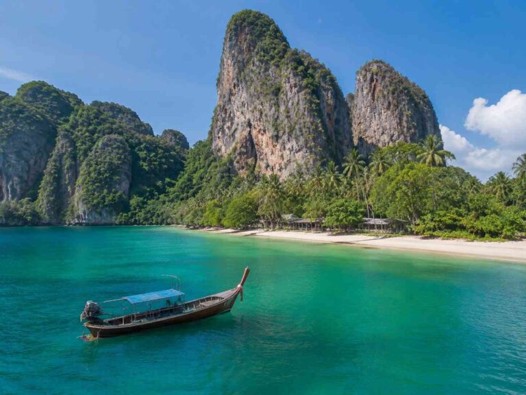 Turquoise sea with a wooden long-tail boat near a white-sand beach, backed by lush green cliffs and palm trees under a blue sky.