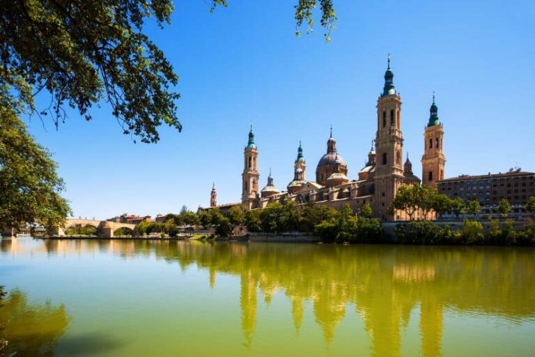 Baroque cathedral with four towers and a central dome by a calm river, trees along the bank, and a blue sky.