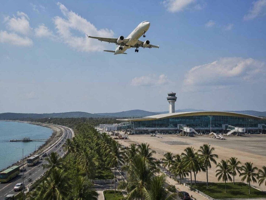 Airplane descending for landing at a seaside airport with a modern terminal and palm trees nearby.