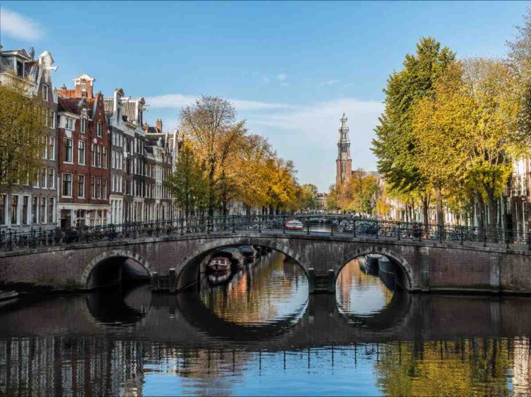 Historic brick arch bridge over a calm canal, with autumn trees and row houses along the water.
