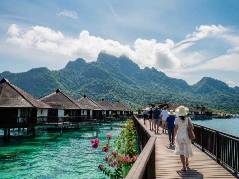 Tourists walk along a wooden boardwalk over turquoise water toward overwater bungalows with a mountain range in the distance under a bright blue sky.