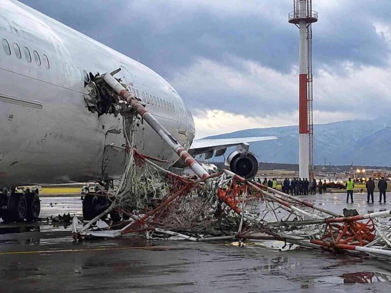 Airliner with a large fuselage breach and tangled, fallen red-and-white support masts on a wet tarmac; emergency personnel in the distance.